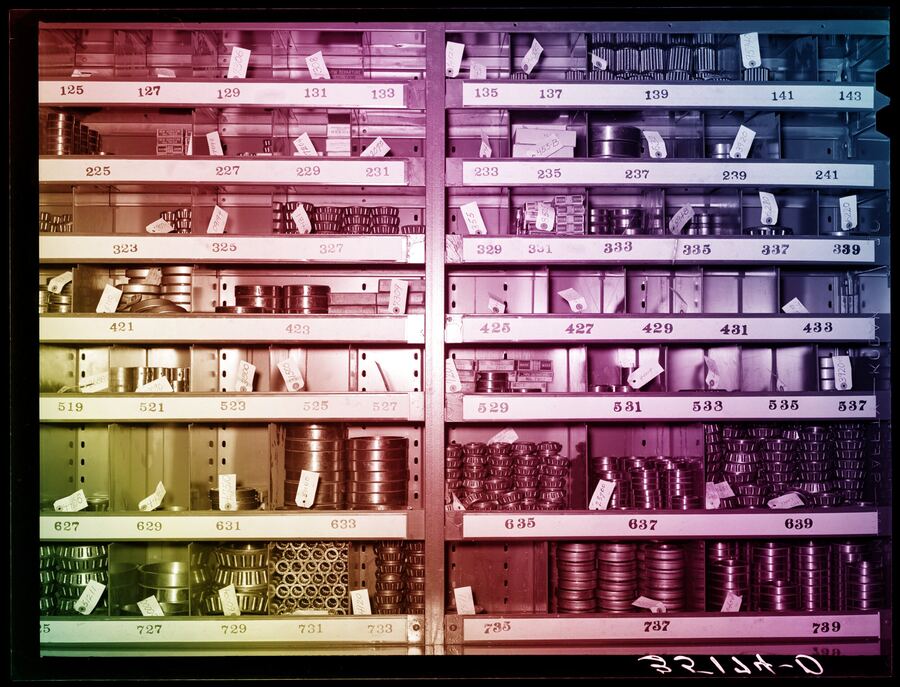 Archival photograph of warehouse shelving divided into numbered bins holding machine parts, tinted with a diagonal rainbow colour cast.