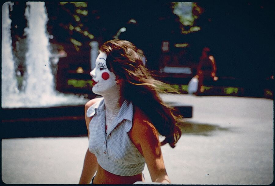 Vintage-toned photo of a young woman in profile with clown-style face paint, a painted winking eye, standing before a plaza fountain.