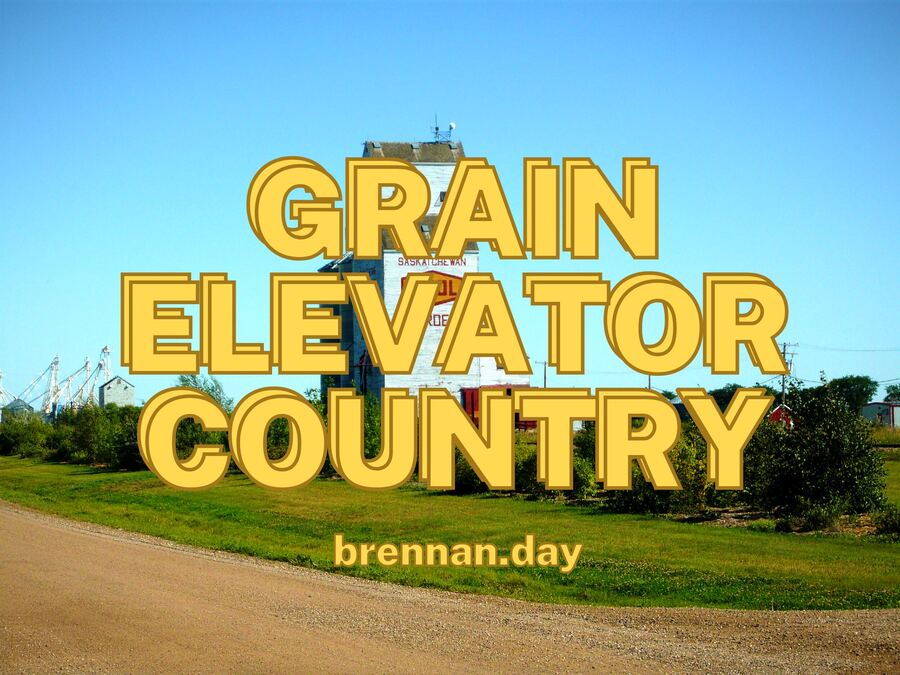 A white wooden grain elevator marked 'SASKATCHEWAN' in a small prairie town under a clear blue sky, with a gravel road running along the foreground.
