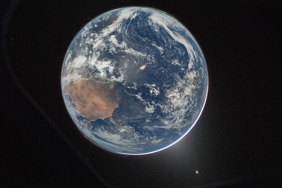 A photograph of Earth from deep space, partially in shadow with a thin luminous blue atmospheric halo, Australia visible as a rust-coloured landmass.