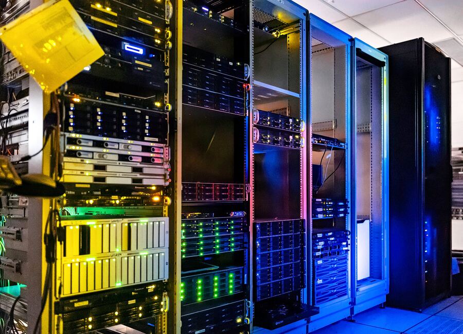A server room with multiple open rack cabinets filled with densely packed hardware, illuminated by dramatic amber, pink, green, and blue lighting.